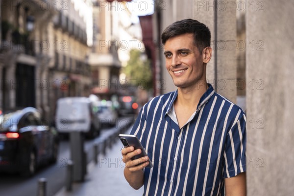 Stylish young man holding a smartphone and smiling on a vibrant city street. Dressed in a striped shirt, he enjoys a relaxed urban moment with a cheerful and modern vibe