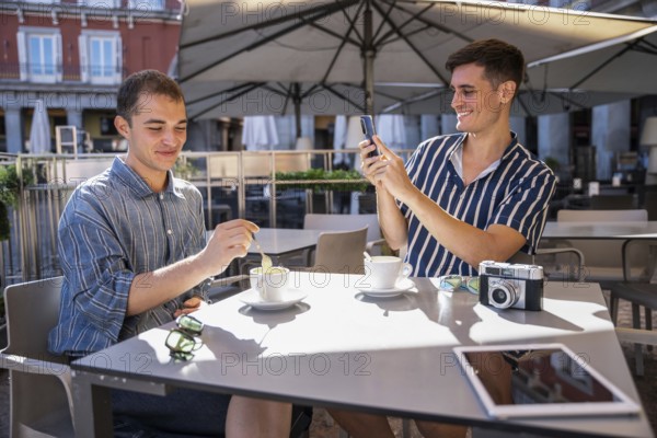 Gay couple enjoying coffee at an outdoor cafe in Plaza Mayor, Madrid. One stirs his coffee while the other captures the moment with a smartphone, blending travel, lifestyle, and relaxation