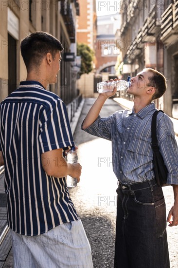 Gay couple pausing for a water break on a sunny street in Madrid. One drinks from a bottle while the other watches, enjoying a casual and relaxed moment in the city