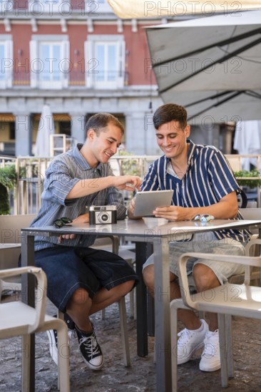 Gay couple sitting at an outdoor cafe in Plaza Mayor, Madrid, smiling while using a tablet and sharing photos from a vintage camera. Relaxed atmosphere with casual fashion and city vibes