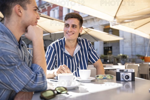 A loving gay couple enjoys a sunny coffee date on the vibrant streets of Madrid, capturing moments of joy and connection. Perfect for themes of love and LGBTQIA+ pride