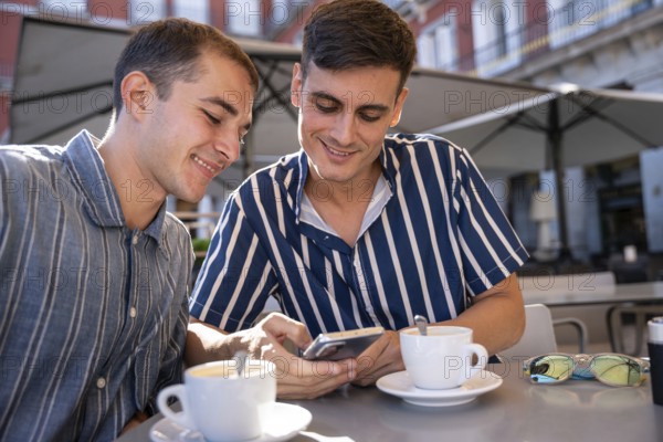 Smiling gay couple enjoying coffee while browsing a smartphone at an outdoor cafe in Plaza Mayor, Madrid. Casual fashion and sunny weather create a relaxed and joyful atmosphere
