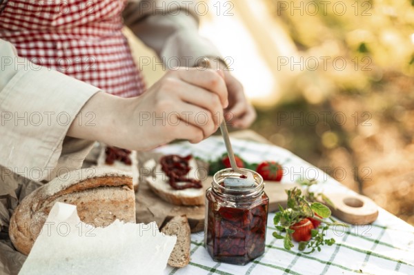 A person prepares canapes using fresh bread, sun-dried tomatoes, and herbs on a picnic table. The vibrant ingredients highlight an inviting culinary scene