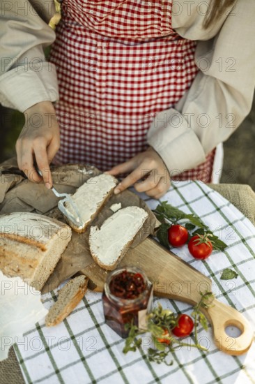 A person wearing a red checkered apron spreads cream on bread slices, surrounded by fresh tomatoes and herbs. The natural setting enhances this culinary scene
