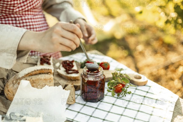 A person spreads sun-dried tomatoes on bread slices for canapes. The setup is outdoors with fresh herbs, creating a rustic and appetizing scene on a gingham cloth