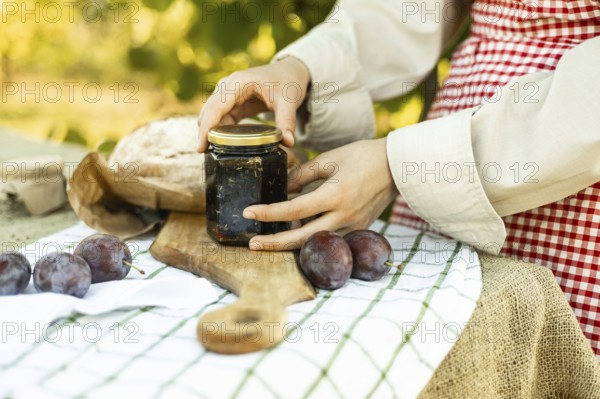 A person prepares canapes using fresh ingredients. The image includes a jar of jam, a loaf of bread, and plums arranged on a cutting board, set on a rustic tablecloth