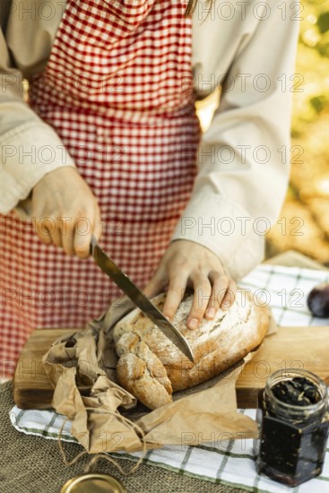 A person slices a loaf of fresh bread on a wooden board, wearing a red check apron. A jar of spread and rustic decor suggest an outdoor canape preparation