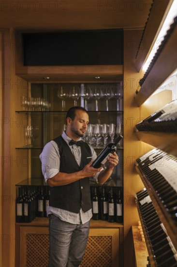 A professional sommelier stands in a cocktail bar, carefully examining a wine bottle. The shelves are filled with bottles and glasses, showcasing an elegant bar atmosphere