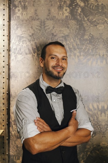 A professional bartender in a stylish cocktail bar setting, dressed in a bow tie and vest, exuding confidence and charm, set against an elegant patterned backdrop