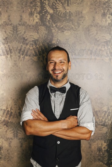 A professional bartender stands confidently with folded arms, wearing a stylish vest and bow tie. The elegant wallpaper backdrop adds to the sophisticated cocktail bar ambiance