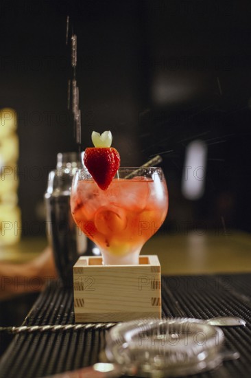 A professional bartender skillfully prepares a vibrant cocktail at a chic bar. The drink is garnished with a fresh strawberry, highlighting a lively atmosphere
