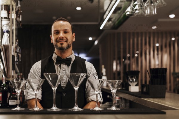 A professional bartender stands confidently behind a stylish bar adorned with martini glasses, exuding expertise and hospitality in a modern cocktail bar setting