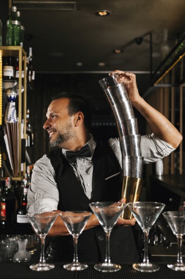 A professional bartender skillfully balances cocktail shakers, showcasing expertise and flair in a stylish cocktail bar. Martini glasses are lined up at the bar, ready for service
