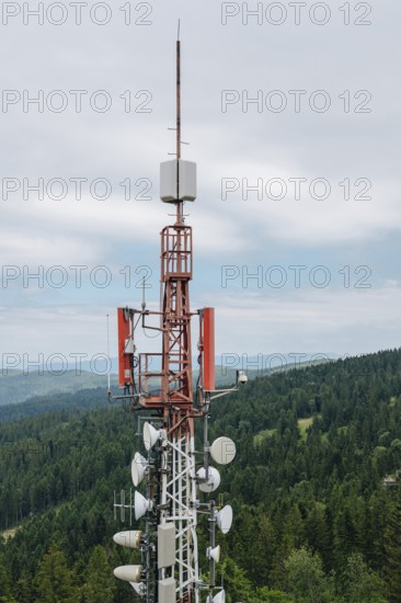 A towering communication array equipped with various antennas and dishes, standing against a backdrop of dense, green forests and distant hills under a cloudy sky. Located near the popular tourist destination of Bachledka in Slovakia