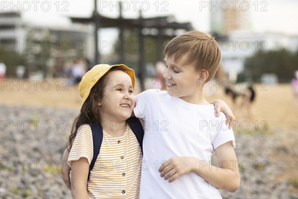 A brother and sister share a joyful moment on a pebble beach. The older boy lovingly wraps his arm around the younger girl. Their smiles echo the warmth of sibling love
