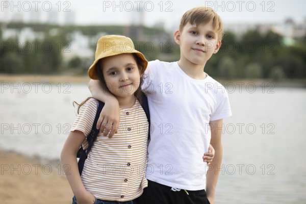 A young brother and sister share a warm moment by a lake. The boy, in a white shirt, lovingly puts his arm around his sister in a striped shirt and yellow hat