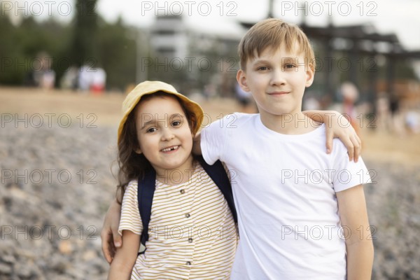 Two smiling siblings embrace while standing on a pebble beach. The young girl wears a sunhat, and the boy in a white shirt enjoys the sunny day. Warm tones evoke joy