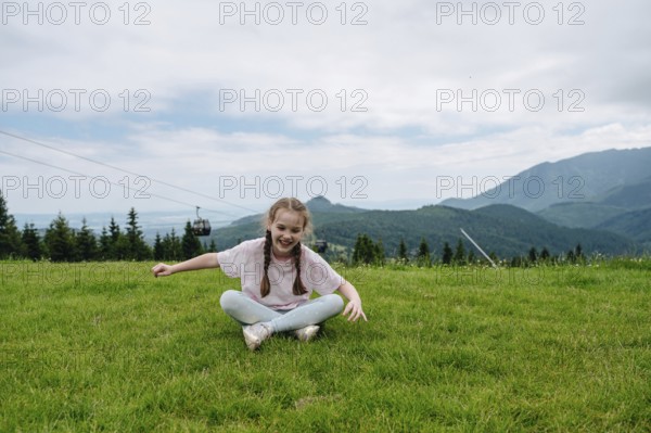 Young girl in casual attire, laughing as she sits on lush green grass at Bachledka Ski Sun in Zdiar, with the Belianske Tatras in the background and a cable car passing overhead