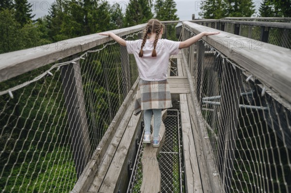 Back view of an unrecognizable girl walking on a treetop bridge at Bachledka. She displays a sense of freedom and exploration while traversing the wooden pathway surrounded by lush foliage