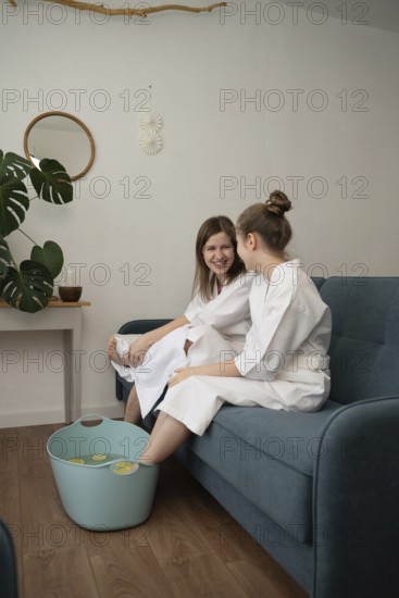 A mother and daughter engage in a self-care routine at home, wearing bathrobes and enjoying a foot soak together on the couch. A bonding moment filled with laughter
