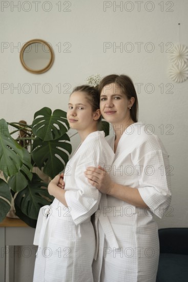 A mother and daughter are enjoying a self-care day at home. They are wearing white bathrobes, standing together, and smiling in a cozy room