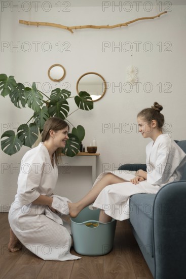 A mother and daughter in cozy bathrobes enjoy a relaxing home spa day. The mother pampers her daughter with a foot bath, fostering bonding and well-being in a cozy, serene space