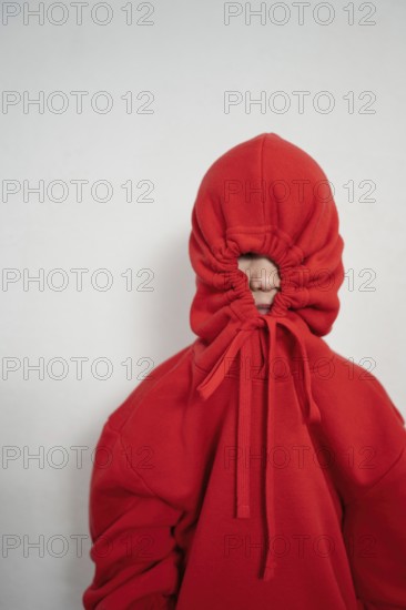 A child wears a red hoodie with the hood pulled tight, completely hiding their face. The drawstrings form a circular pattern around the small opening