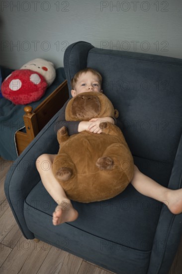 A child relaxes in a cozy chair, cuddling a large plush bear. The scene exudes comfort and warmth, with soft lighting and a playful atmosphere in the background