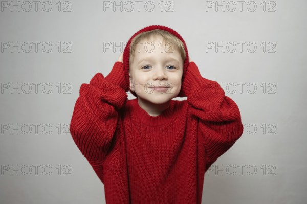 A cheerful young child wearing an oversized red sweater and hat, smiling playfully at the camera. The vibrant color and cozy texture create a warm, joyful atmosphere