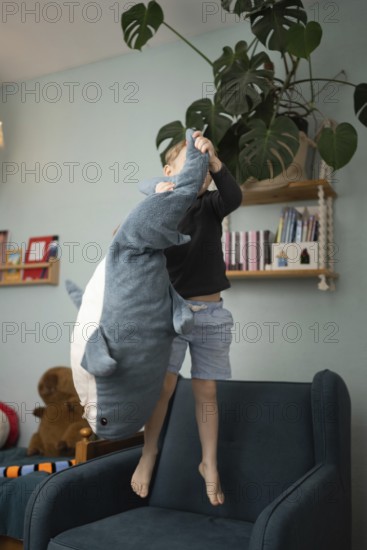 A child joyfully playing with a large shark plush toy while standing on a chair in a cozy, well decorated room with books and plants