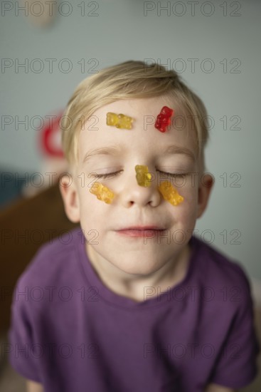 A child with closed eyes smiles blissfully with colorful gummy bears on their face, expressing a playful and humorous moment. The child wears a purple shirt