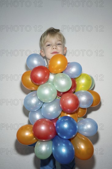 A cheerful child is standing with a delightful expression, almost hidden behind a vibrant bunch of colorful balloons. The scene exudes joy and playfulness in a simple background