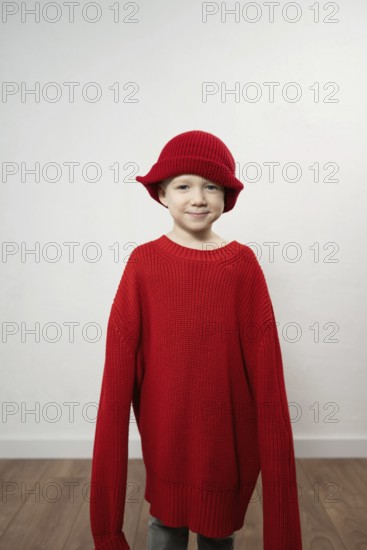 A child wears an oversized red sweater and matching hat, smiling playfully. The neutral background highlights the vibrant clothing, emphasizing a whimsical and cozy theme