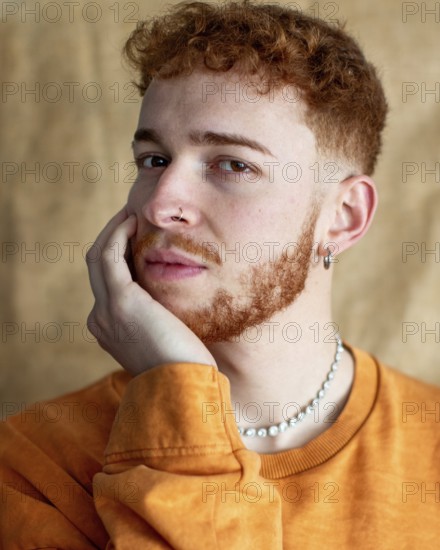 A close up portrait of a stylish young man with red curly hair, wearing a pearl necklace and earrings. He gazes thoughtfully, with his hand on his cheek, in casual attire