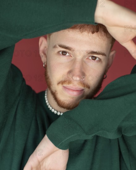 A man red hair wearing a green sweater poses with arms creatively framing their face against a red backdrop. The expressive facial expression is highlighted by subtle jewelry