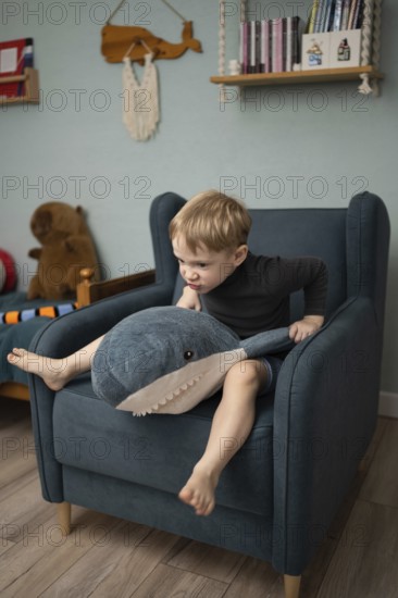 A child sits on an armchair, energetically interacting with a plush shark toy. The cozy room features books, wall decor, and stuffed animals, creating a playful atmosphere