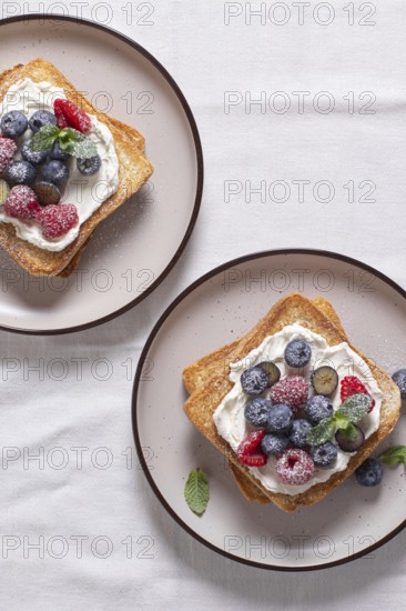 Top view of two toasts with cream cheese topped with raspberries, blueberries, and dusted with powdered sugar, served on ceramic plates