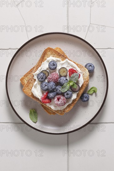 Top view of a delicious toast topped with cream cheese, raspberries, blueberries, and a sprinkle of powdered sugar, served on a dark rimmed plate
