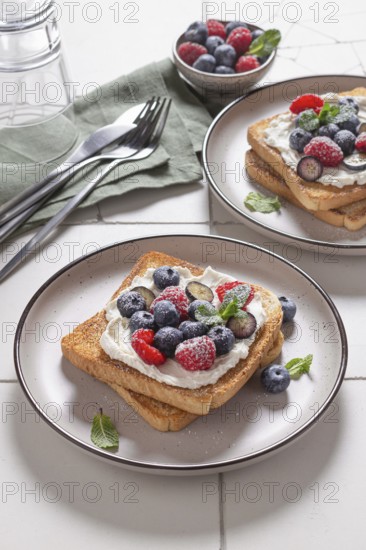 Plated serving of toast layered with cream cheese and topped with an assortment of fresh berries such as blueberries and raspberries, garnished with mint leaves, on a speckled plate beside silverware
