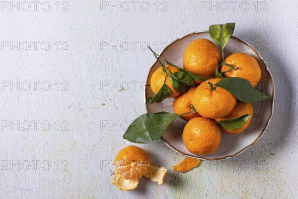 A bowl of fresh tangerines with green leaves, arranged on a textured surface. One tangerine is partially peeled, highlighting its vibrant orange color and juicy texture