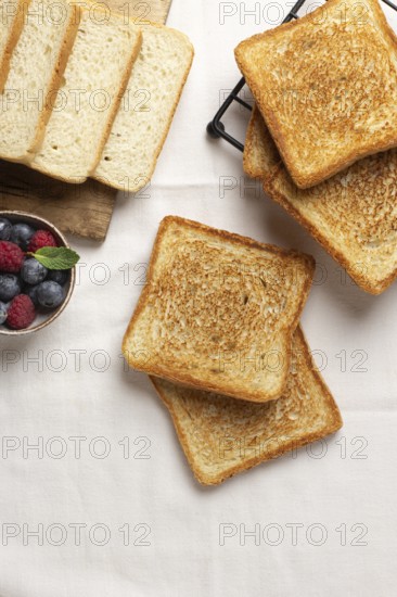 Top view of golden toasts alongside a bowl of blueberries and raspberries on a rustic wooden board and cloth background