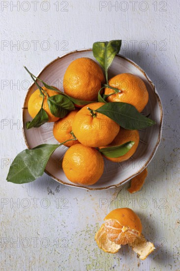 A vibrant arrangement of fresh tangerines with green leaves, displayed on a rustic plate against a textured background. One tangerine is peeled, revealing the juicy segments