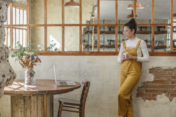 A young woman, holding a coffee cup, engages in telework at a rustic cafe table with her laptop open next to a decorative vase