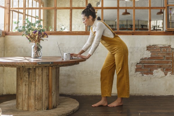 A young woman, dressed in yellow overalls, is focused on her laptop at a wooden circular table in a room with rustic decor, symbolizing a home office environment