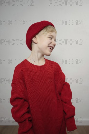 A joyful child wearing an oversized red sweater and matching beanie, smiling widely. The plain background accentuates the vibrant attire and lively expression