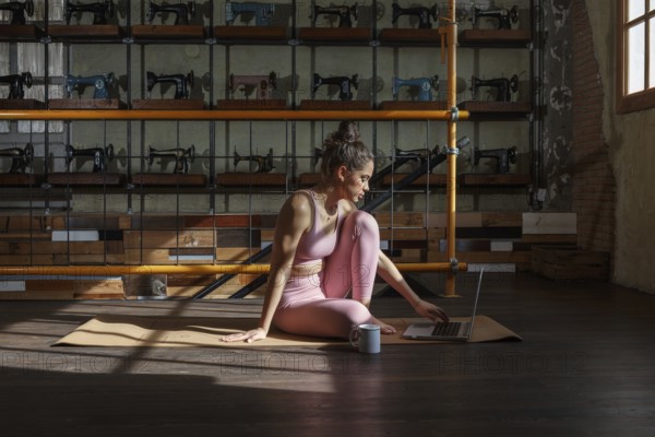 Woman in athletic attire practices yoga on a mat while working on a laptop. Sunlight filters through a window, creating a cozy and productive atmosphere
