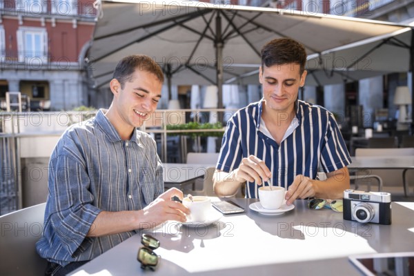 A gay couple enjoys a sunny day while having coffee at a cafe on the streets of Madrid. The scene captures a moment of happiness and companionship
