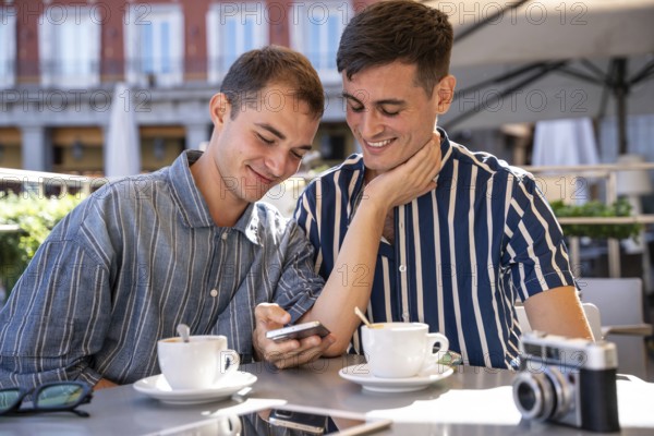 A gay couple shares a joyful moment at an outdoor cafe in Madrid. They smile while looking at a phone, with coffee and a camera nearby, capturing a casual, intimate scene
