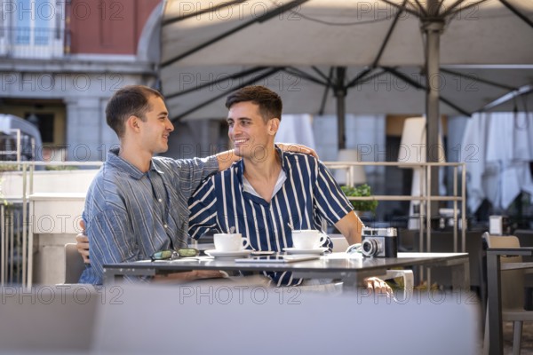 A gay couple shares a joyful moment at a charming outdoor cafe on the streets of Madrid. They are seated at a table, enjoying each other's company in a sunny ambiance