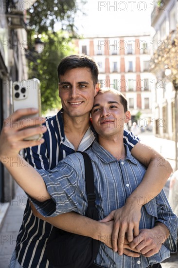 A gay couple embraces while taking a selfie on a picturesque street in Madrid. Sunlight filters through nearby trees, capturing a vibrant and joyful moment